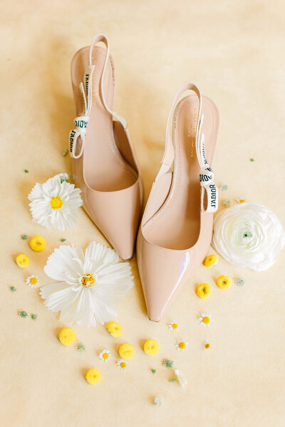 Taupe high heels with a pointed toe and thin heel strap set on a neutral backdrop with white and yellow petals