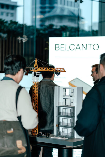A building design with a toy crane on a table with people looking at it with Belcanto words in the background