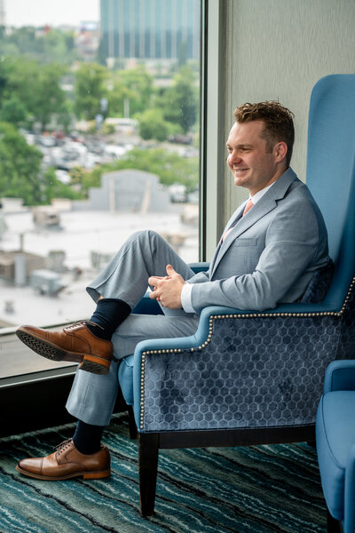 Groom sitting in blue armchair adjusting his tie and smiling confidently – photographed by wedding photographer Niko Coric – Lumen Clarity Media