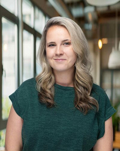 A person stands on stone steps in front of a large stone building, holding a bright red coat. They are wearing a white dress and red shoes. Trees are visible in the background.
