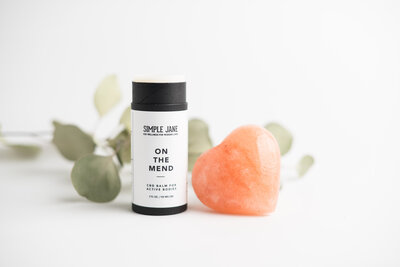 CBD Balm stick sitting on a white table with Eucalyptus and heart shaped salt stone
