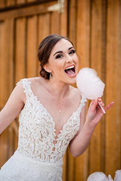 Bride wearing a form fitting lace dress eating cotton candy and smiling at the camera