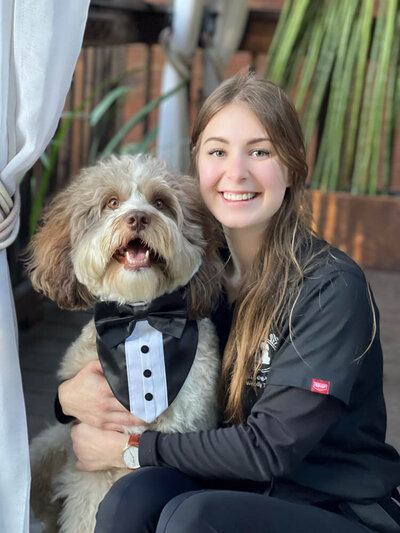 woman with black shirt with a fluffly brow/grey dog next to her wearing a black tie-on tuxedo