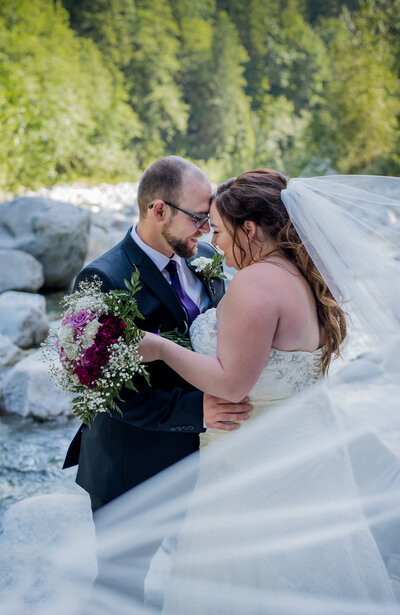 Bride and groom foreheads touching , river in the back round and veil coming to the side.