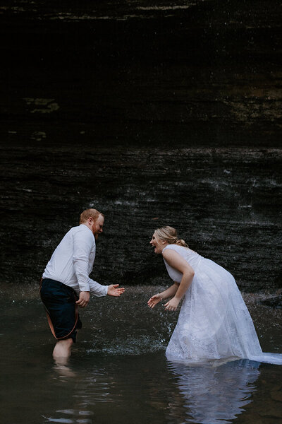An eloping couple in wedding attire is knee-deep in a pool of water created by Bridal Veil Falls in Heber Springs, Arkansas. They are splashing water at each other and laughing.
