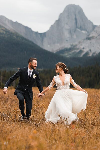 Stunning mountain backdrop for gorgeous calgary wedding, photographer captures bride and groom running through a field.