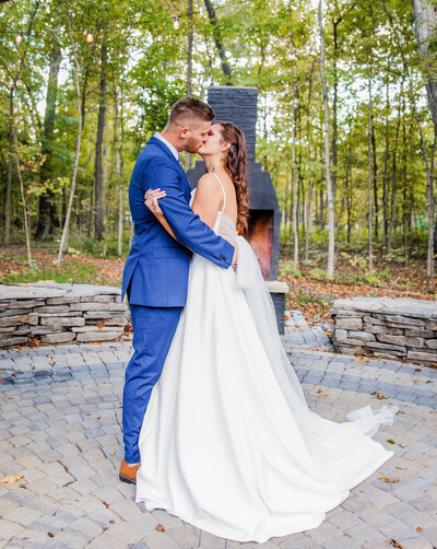 A bride and groom kiss each other in a forest that is full of fall trees. He is wearing a blue suit and she is wearing a long white dress. They are standing in front of a firepit.