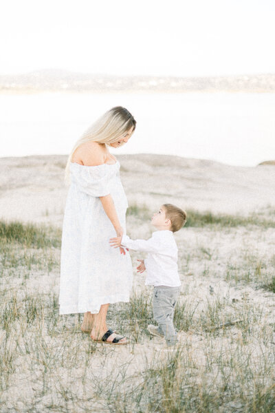 Image of expecting mother in white dress standing at the lake holding her sons hand and looking at eachother taken by Sacramento Maternity Photographer Kelsey Krall