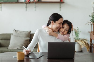 Mother sitting at a desk working on her tablet and laptop while her daughter hugs her neck.