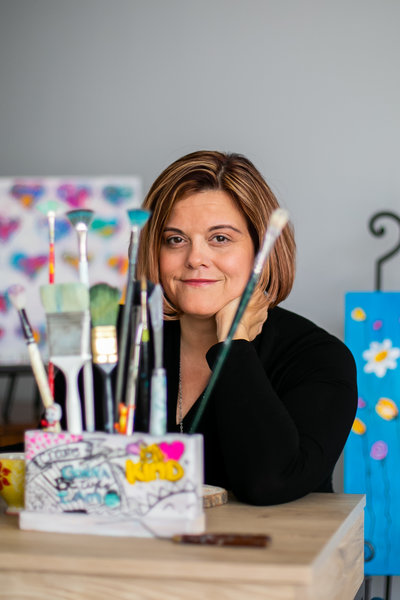 Artist in black sweater poses for photo behind her paint brushes in her studio