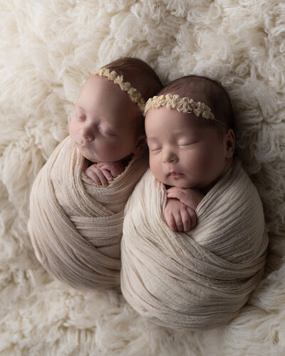 Sleeping twin newborns wearing cream wraps and on a cream colored rug with bow headbands