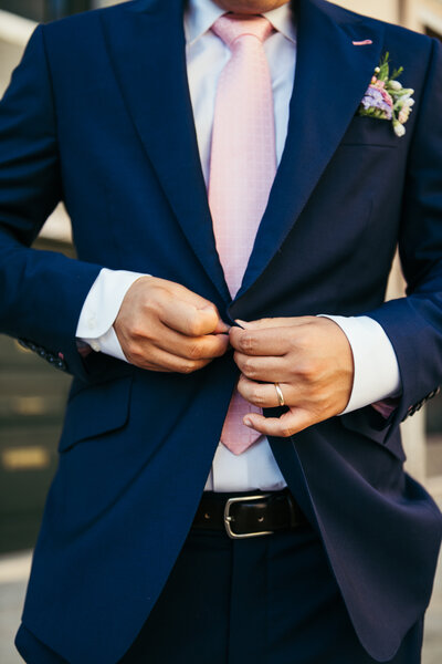 A close-up captures the groom's hands skillfully buttoning his jacket, showcasing the meticulous details of his attire in anticipation of the wedding ceremony