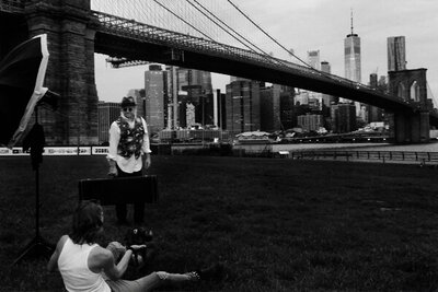 black and white bts image Mark Maryanovich sitting on ground photographing musician Robert Allen standing by Brooklyn Bridge flash directed at him