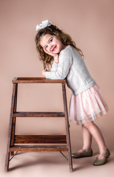 A little girl with a big bow in her hair is leaning on a wooden ladder and smiling at her studio session  with Ashley Zohil Photography.