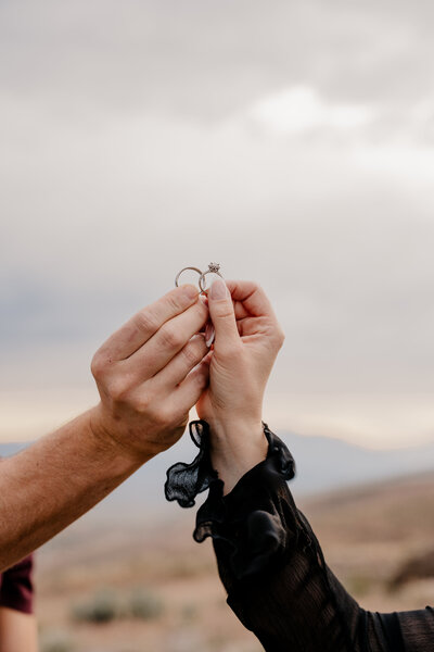 The couple holds their wedding rings up in the sky, with the clouds as the background.