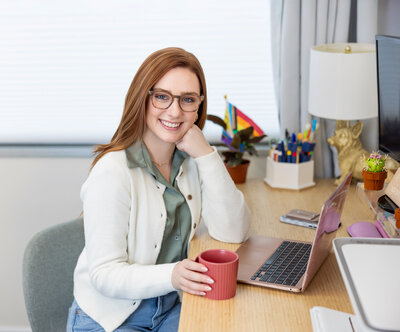 A-therapist-smiles-with-laptop-and-lgbtqia+-flag-in-office-during-brand-photoshoot-in-Baltimore