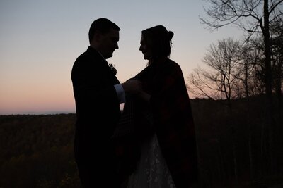 a groom helps his bride adjust a shawl as their luxury destination elopement photographer captures them silhouetted against the orange, yellow and blue ombre sunset sky.  