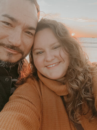 Woman in burnt orange sweater and man in black with beard take a selfie on the beach in Southern California at sunset