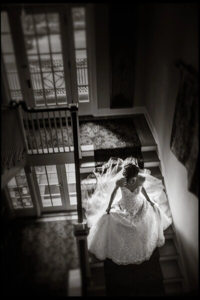 Black and white image of a bride in a lace gown looking downwards as she descends a staircase with natural light streaming through the window.