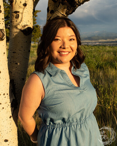 photo of Ashlyn in a blue denim dress standing with her back against an aspen tree