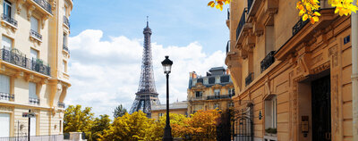 A view of the Eiffel Tower in Paris, framed by classic Parisian buildings under a bright blue sky. Autumn leaves in shades of yellow and orange line the street, adding a touch of warmth to the scene.