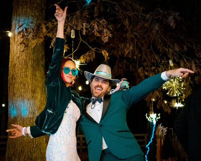 A wedding couple with colorful sunglasses and hats on celebrating and holding their arms out.