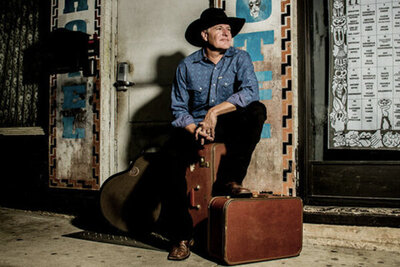 Country musician portrait Darrell Goldman sitting on suitcase one foot resting on another piece of luggage guitar resting on wall behind him