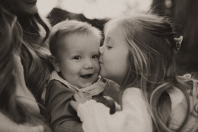 Big sister kisses her littler brother on the cheek in black and white.
