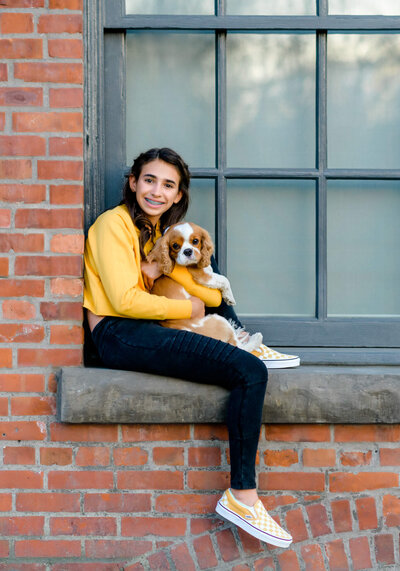 A girl in a yellow sweater sits in a windowsill holding her small dog