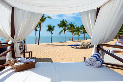 View from a beach cabana with white curtains, overlooking sandy beach and palm trees. Rolled blue and white towels are on the cabana bed. Sun loungers face the calm blue sea under a clear sky.