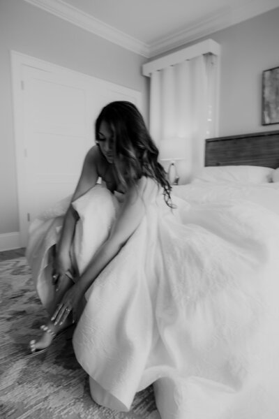 Bride photographed sitting on bed and putting on shoes
