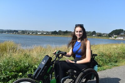 Bethany Handley smiling outdoors in a wheelchair on a lakeside path, wearing a blue top on a sunny day.