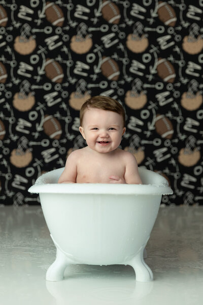 A toddler boy sits in a tiny bathtub in a football themed New Jersey Milestone Photographer studio