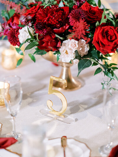 Chic gold acrylic table numbers backed by vibrant red florals in a gold vase at a December wedding reception at the Don Cesar Hotel, St. Pete Beach, FL.