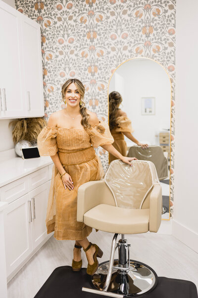 woman standing next to hair stylist chair wearing a loose dress and smiling