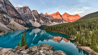A serene lake reflects the surrounding tall, rugged mountains at sunset, with their peaks tinted orange by the sun. The calm blue water is bordered by dense green pine forests. The sky is clear with a hint of dusk light.