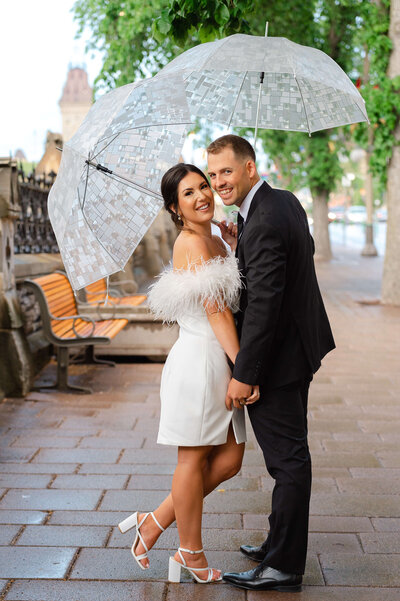 a bride in a short white dress smiles with her groom in a black suit under their umbrellas on Wellington street taken by Ottawa wedding photographer JEMMAN Photography