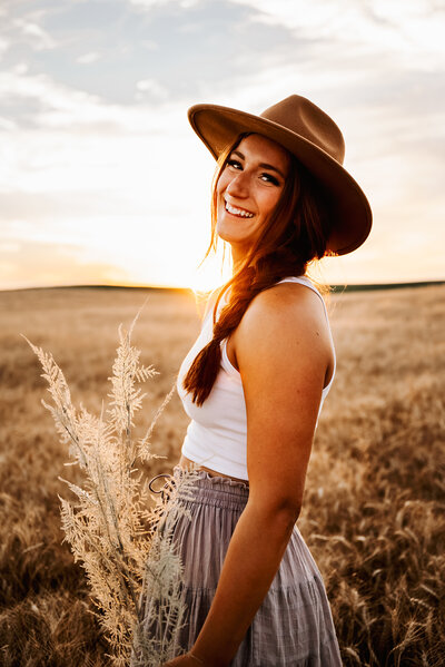 Girl laughs as she holds plant and stands in golden wheat field at sunset