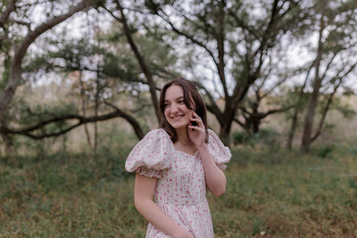 A young highschool senior girl smiling with her hand lightly touching her face looking off into the distance in a field