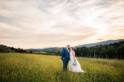 Shenandoah wedding photographed by charlottesville wedding photographer with bride adn groom in a yellow wildflower field leaning in to kiss each other as the sunsets over the mountains in the distance for a summer wedding