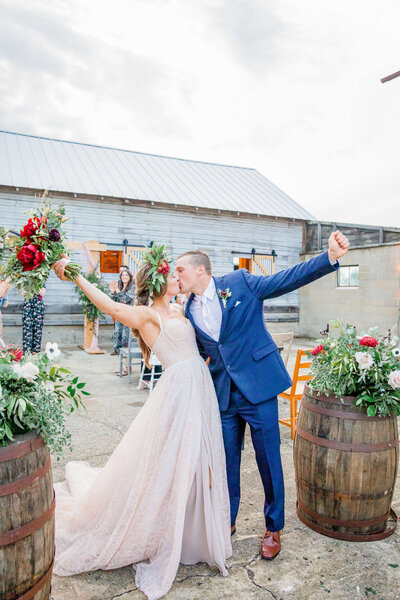 A bride and groom, who have just gotten married, are at the end of their wedding aisle. They have their hands up in celebration and kiss while raising the bouquet. There are two barrells next to them.
