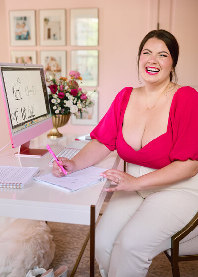 Brunette woman wearing hot pink shirt, working at her desk, smiling at the camera