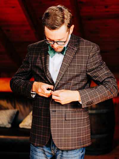 This image features a groom wearing a plaid suit jacket and a green bowtie. The groom is buttoning his jacket while standing in his dressing room at the wedding venue. This image was taken by KD Captures, a wedding photographer in San Antonio.