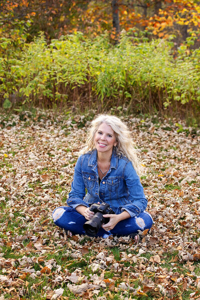 Female photographer sitting on the ground outdoors on colorful fall leaves holding camera.