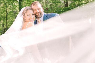 A happy bride and groom during their portraits on a spring wedding day in Raleigh with the brides wedding veil blowing in the wind by JoLynn Photography, a Raleigh wedding photographer