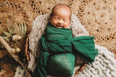a baby wrapped in a green wrap laying in a basket for their newborn photo shoot