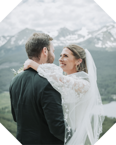 Grinning bride in long-sleeved wedding dress and veil grinning at groom in mountain landscape setting - professional photo edit