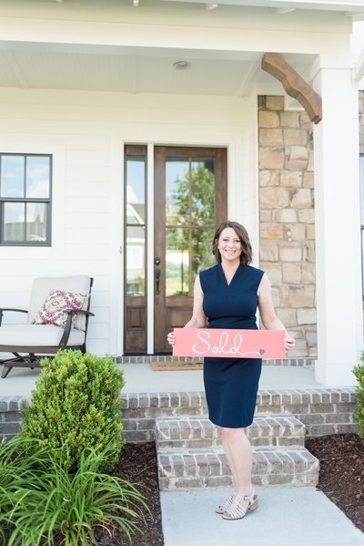 Nashville realtor wearing a navy dress posing in front of a house with a red sign that says "sold"
