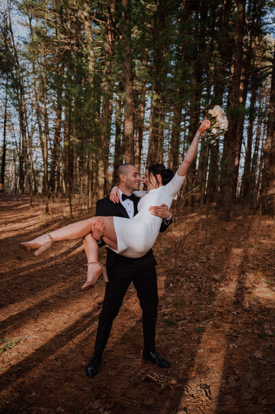 Groom holding bride in his arms while bride holds bouquet up and smiling while in the woods