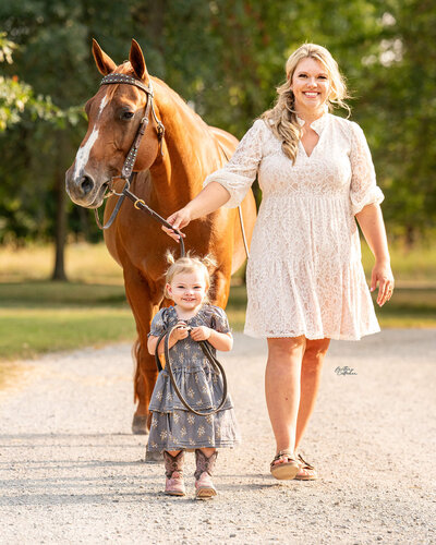 Blonde equestrian wearing a cream dress with her daughter with pigtails and a blue dress walking down a gravel pathway with their chestnut chestnut quarter horse with green trees behind them.